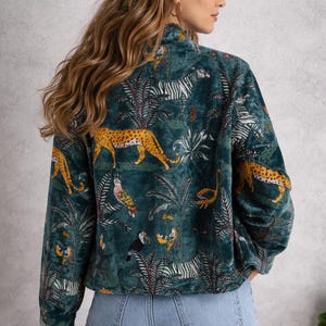 May include: Teal velvet jacket with a jungle-themed print. The design features zebras, leopards, birds, and tropical foliage. The jacket has a high collar and long sleeves, and is paired with blue jeans.