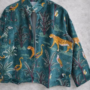 May include: Teal velvet jacket with a jungle-themed print. The jacket features a repeating pattern of leopards, zebras, birds, and palm trees. The interior lining is striped. The jacket is hanging on a wooden hanger.