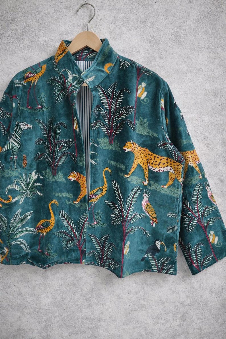 May include: Teal velvet jacket with a jungle-themed print. The jacket features a repeating pattern of leopards, zebras, birds, and palm trees. The interior lining is striped. The jacket is hanging on a wooden hanger.