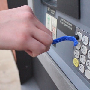 May include: A blue "Kooly Key" tool is used to press the number 2 on an ATM keypad. The tool has a curved design with the text "Kooly Key" printed on it. The ATM keypad has white buttons with black numbers and letters.