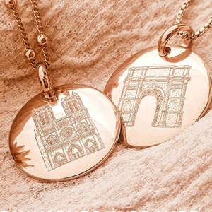 May include: Two gold necklaces with round pendants featuring engraved images of famous Parisian landmarks: the Arc de Triomphe and Notre Dame Cathedral. The text "MONUMENTS À GRAVER" and the names of the landmarks are visible below the pendants.