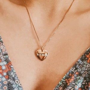 May include: A gold heart-shaped pendant necklace with a delicate chain. The pendant has an inscription on the front.