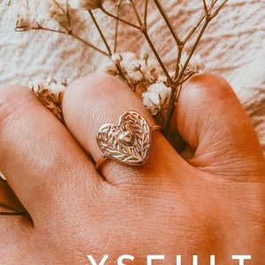 May include: A gold heart-shaped ring with a detailed leafy design. The ring is worn on a finger and the background is blurred.