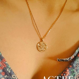 May include: A gold chain necklace with a round pendant featuring a detailed floral design. The pendant is hanging on a person's chest. The text "ACTÉE" is visible at the bottom of the image.