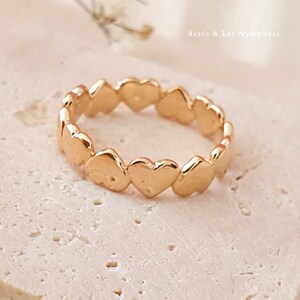 May include: Gold ring with a band of connected heart shapes. The ring is on a light beige surface.