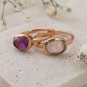 May include: Two gold rings with oval shaped gemstones. The ring on the left has a purple amethyst stone. The ring on the right has a light pink rose quartz stone.