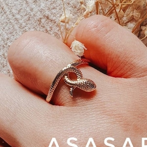 May include: A gold snake ring with a textured finish. The ring is worn on a person's finger. The text "ASASARA" is visible in the image.