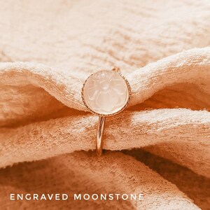 May include: A gold ring with a white moonstone set in a delicate floral design. The ring is on a light brown fabric background.  ENGRAVED MOONSTONE