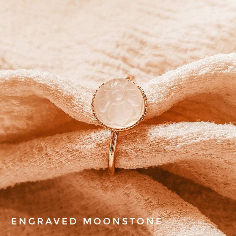 May include: A gold ring with a white moonstone set in a delicate floral design. The ring is on a light brown fabric background.  ENGRAVED MOONSTONE