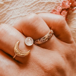 May include: Close-up of three gold-colored rings on a hand. One ring features a crescent moon design, another has a round gemstone, and the third has a floral pattern. The word "ACT&Eacute;E" is visible at the bottom.