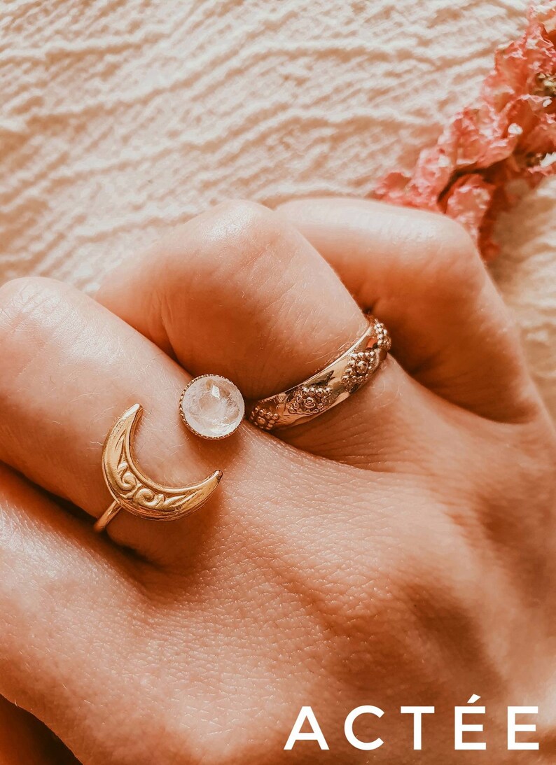 May include: Close-up of three gold-colored rings on a hand. One ring features a crescent moon design, another has a round gemstone, and the third has a floral pattern. The word "ACT&Eacute;E" is visible at the bottom.
