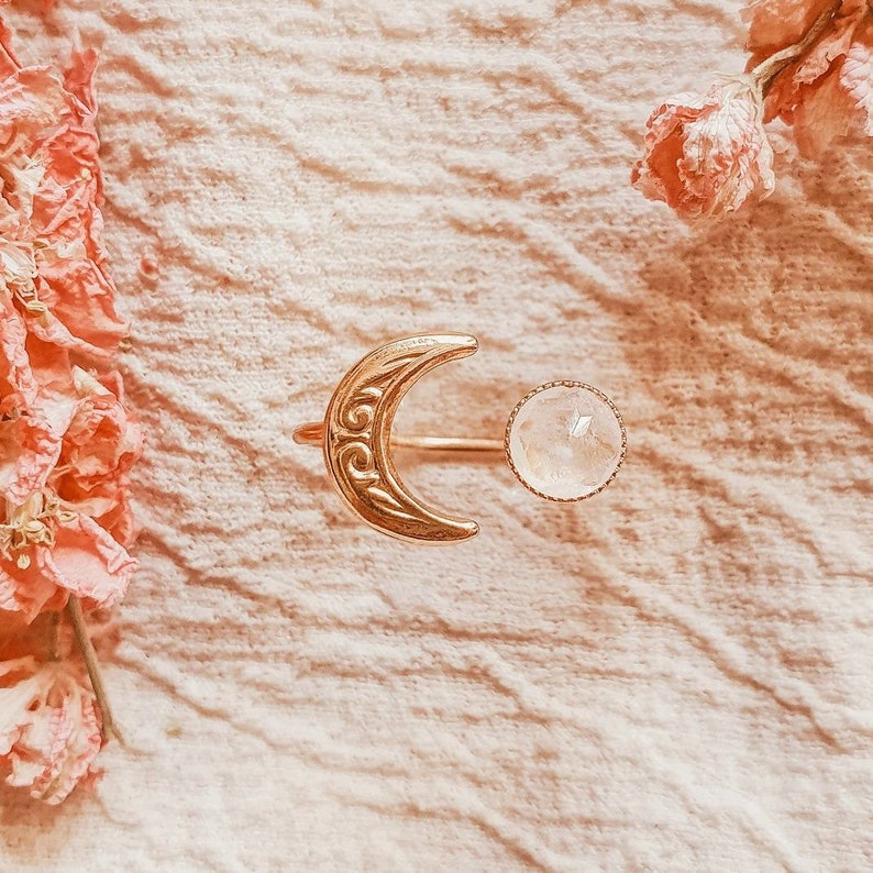 May include: A gold-colored ring featuring a crescent moon design with intricate scrollwork and a round, clear gemstone. The ring is set against a soft, textured background with pink floral accents, creating a delicate and celestial aesthetic.