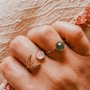 May include: Three rings are displayed on a hand. One ring features a gold crescent moon design. Another ring has a clear, round stone. The third ring has a green, round stone. The rings are gold-toned.