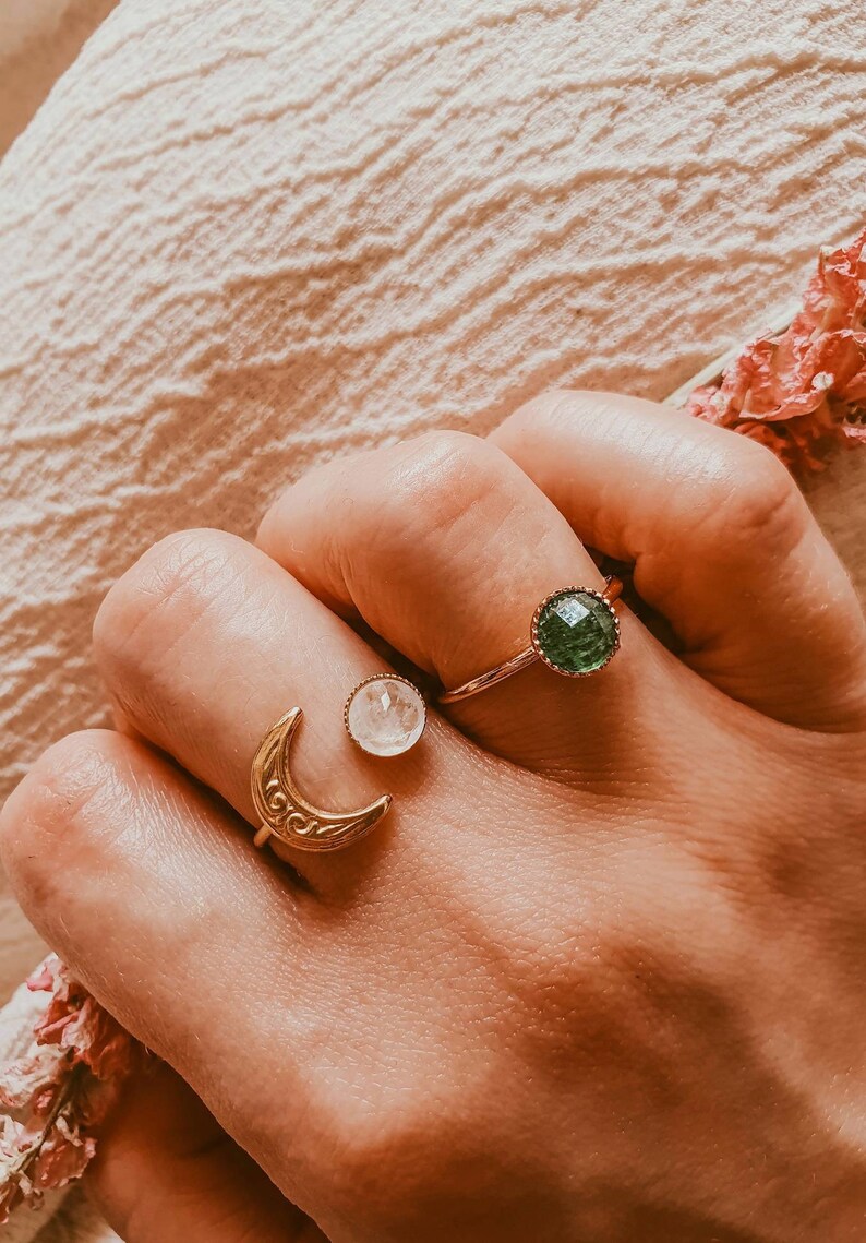 May include: Three rings are displayed on a hand. One ring features a gold crescent moon design. Another ring has a clear, round stone. The third ring has a green, round stone. The rings are gold-toned.