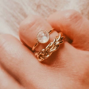 May include: Two gold rings on a finger. The top ring features a clear, faceted gemstone. The bottom ring is a braided design.