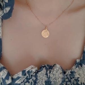 May include: A gold chain necklace with a round pendant featuring a detailed image of a religious figure. The necklace is worn by a person wearing a blue and white floral patterned shirt.