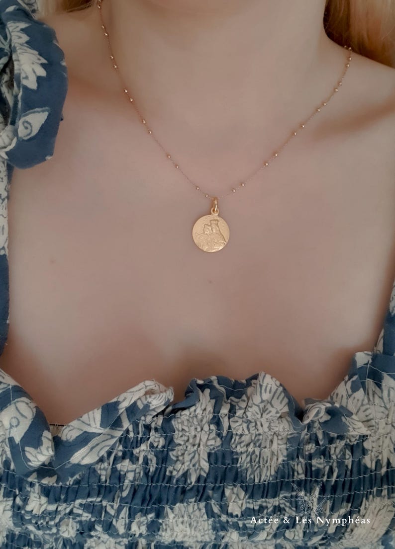 May include: A gold chain necklace with a round pendant featuring a detailed image of a religious figure. The necklace is worn by a person wearing a blue and white floral patterned shirt.