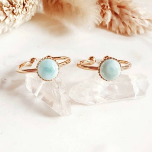 May include: Two gold rings with a light blue gemstone set in each. The rings are on a white surface with a clear crystal next to each ring.
