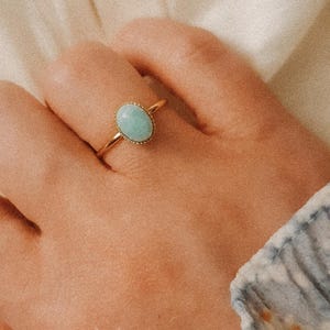 以下が含まれることがあります： 薄い青色の楕円形の宝石が付いた金の指輪。指輪は人の指にはめられています。