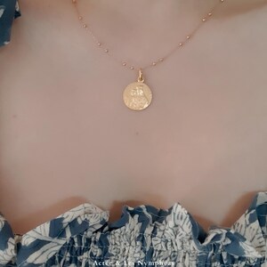 May include: A gold chain necklace with a round pendant featuring a detailed design. The necklace is worn by a person with a blue and white floral patterned shirt.