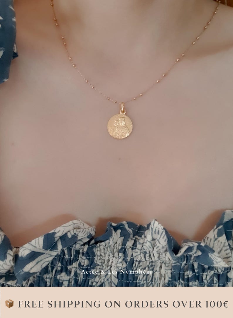 May include: A gold chain necklace with a round pendant featuring a detailed design. The necklace is worn by a person with a blue and white floral patterned shirt.