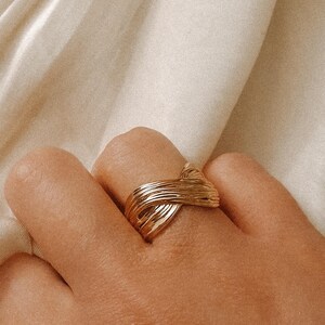 May include: A gold-toned ring with a woven design. The ring is worn on a finger.