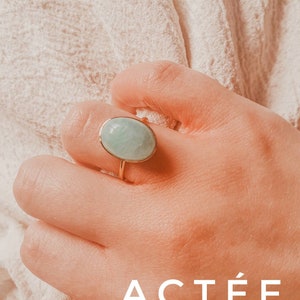 May include: A gold ring with a light blue oval gemstone. The ring is on a person's finger. The text "ACTÉE" is visible in the image.