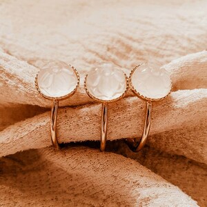 May include: Three gold napkin rings with clear, white, round, carved glass stones. The rings are on a beige fabric surface.
