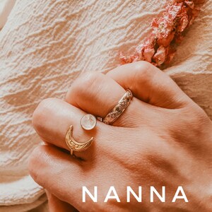 May include: A hand wearing three rings: a gold crescent moon ring, a small round ring with a white stone, and a textured band. The word "NANNA" is displayed in white block letters at the bottom of the image. Pink dried flowers are in the background.
