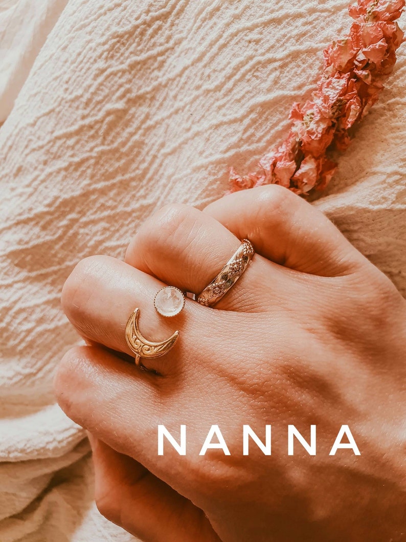 May include: A hand wearing three rings: a gold crescent moon ring, a small round ring with a white stone, and a textured band. The word "NANNA" is displayed in white block letters at the bottom of the image. Pink dried flowers are in the background.