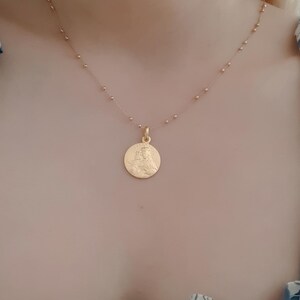 May include: A gold chain necklace with a round pendant featuring a detailed image of a woman. The chain is made of small, evenly spaced beads.