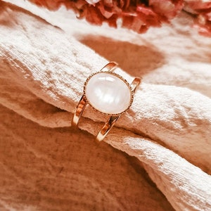 May include: A gold ring with a white oval gemstone set in a bezel setting. The ring is on a white fabric background.