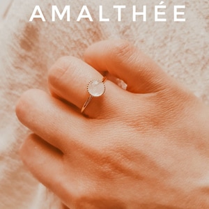 May include: A gold ring with a round, light-colored stone is worn on a finger. The ring has a delicate, textured setting. The word "AMALTHÉE" is displayed in white capital letters above the ring. The background is a soft, neutral color.