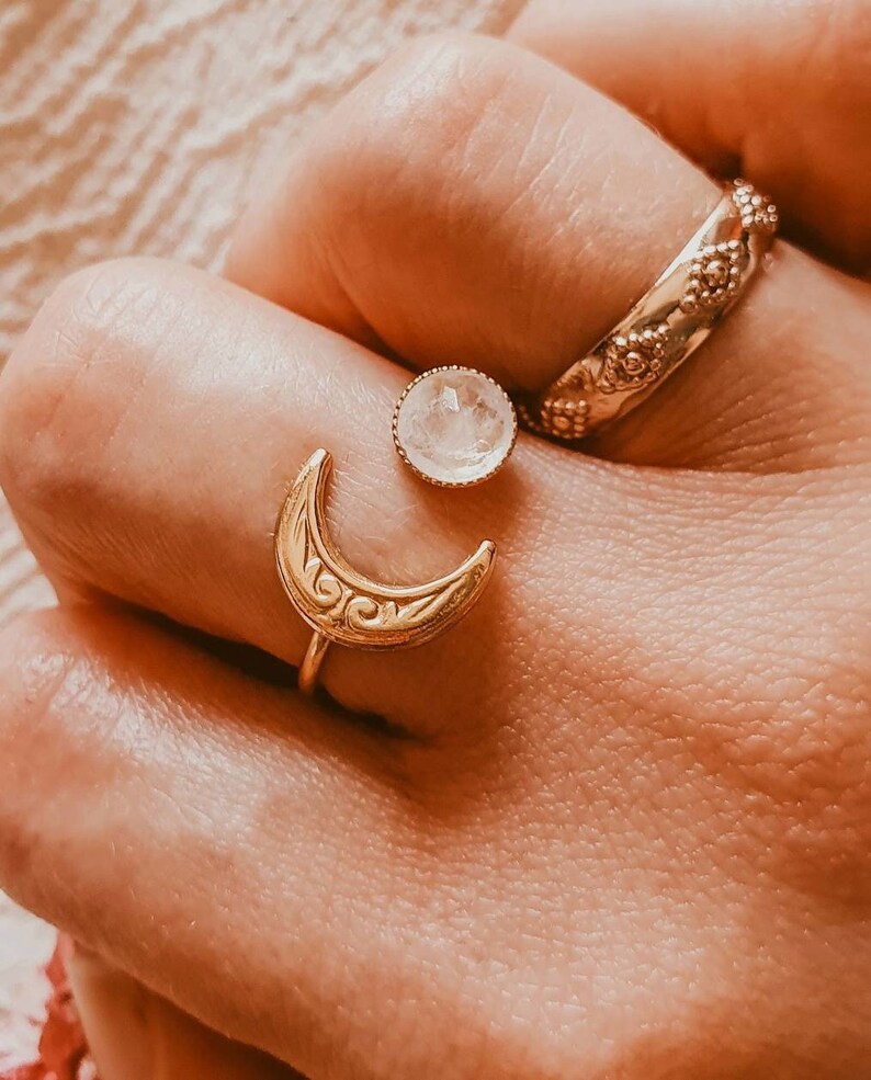 May include: A hand displays three gold rings. One ring is a crescent moon shape with an ornate design. Another is a round ring with a floral pattern. The third ring is a small, round, clear gemstone set in a gold band.