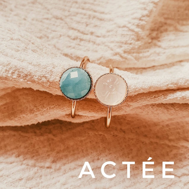 May include: Two gold rings with a blue and a white stone. The blue stone is a light blue colour and the white stone is a milky white colour. The rings are on a light brown fabric background. The word "ACT&Eacute;E" is written in white letters on the bottom of the image.