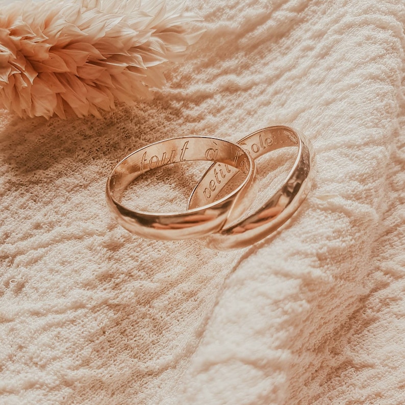 Pu&ograve; includere: Due fedi nuziali d'oro con le parole "tout" e "soleil" incise. Le fedi sono appoggiate su una superficie di tessuto beige.