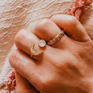 May include: A hand wearing three gold rings. One ring features a crescent moon design, another has a round, clear stone, and the third is a band with an ornate pattern. A sprig of pink flowers is in the background.