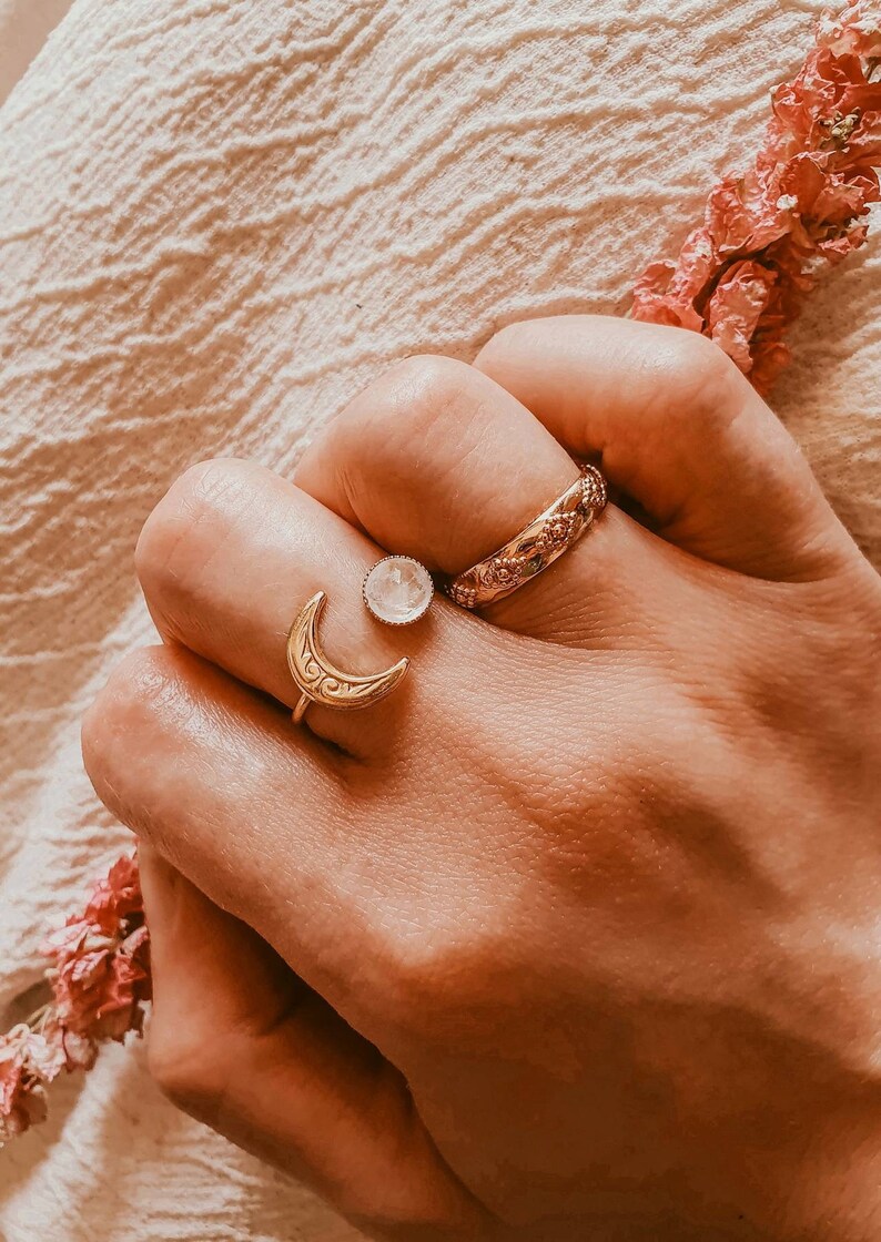May include: A hand wearing three gold rings. One ring features a crescent moon design, another has a round, clear stone, and the third is a band with an ornate pattern. A sprig of pink flowers is in the background.