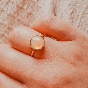 May include: A gold ring with a white opal gemstone set in a bezel setting. The ring is on a person's finger.