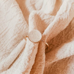 May include: A gold ring with a white, round, carved stone. The ring is on a white fabric background.
