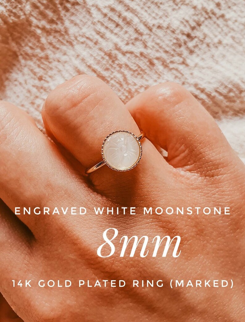 May include: A gold plated ring featuring an 8mm white moonstone with an engraved floral design. The ring is marked 14k.