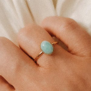 以下が含まれることがあります： 薄い青色の楕円形の宝石が付いた金の指輪。指輪は人の指にはめられています。
