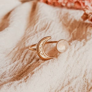 May include: A gold-colored ring featuring a crescent moon design with etched details and a round, light-colored stone. The ring is set against a soft, textured background, creating a delicate and celestial aesthetic. The ring's design is open, with the moon and stone separated.