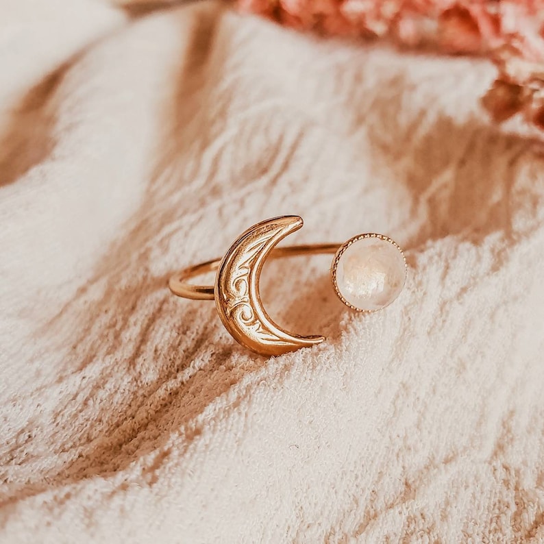May include: A gold-colored ring featuring a crescent moon design with etched details and a round, light-colored stone. The ring is set against a soft, textured background, creating a delicate and celestial aesthetic. The ring's design is open, with the moon and stone separated.