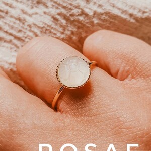 May include: A gold ring featuring a white, round, carved stone. The stone has a delicate design and is set in a gold bezel. The ring is on a finger.