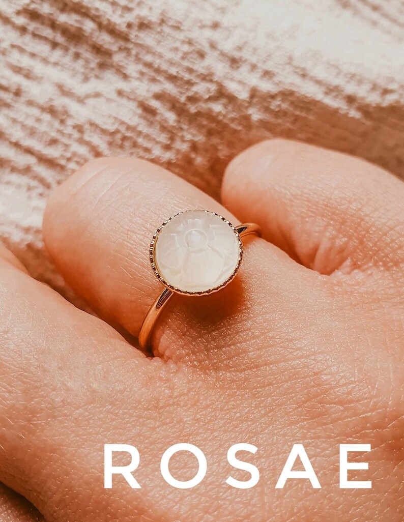 May include: A gold ring featuring a white, round, carved stone. The stone has a delicate design and is set in a gold bezel. The ring is on a finger.