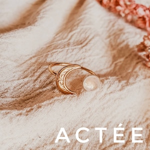 May include: Gold-toned ring featuring a crescent moon design with etched details and a round, light-colored stone. The ring is open-ended and sits on a textured, off-white surface. The word "ACT&Eacute;E" is displayed in white text at the bottom.