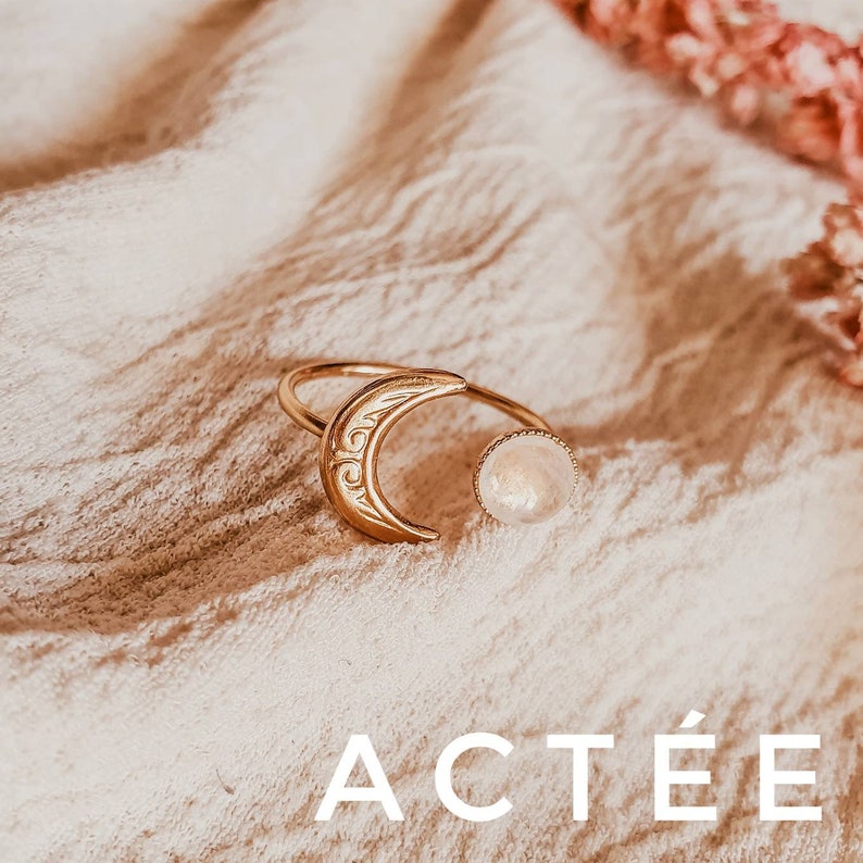 May include: Gold-toned ring featuring a crescent moon design with etched details and a round, light-colored stone. The ring is open-ended and sits on a textured, off-white surface. The word "ACT&Eacute;E" is displayed in white text at the bottom.