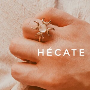May include: A gold-colored ring with a central clear stone and crescent moon accents. The word "HÉCATE" is displayed in white text. The ring is worn on a finger.