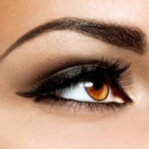 以下が含まれることがあります： 茶色の虹彩、濃いアイライナー、長く濃いまつげを持つ女性の目のクローズアップ。目は、よく定義されたアーチ型の眉によって縁取られています。目の周りの肌は滑らかで明るい色調で、微妙な陰影があります。